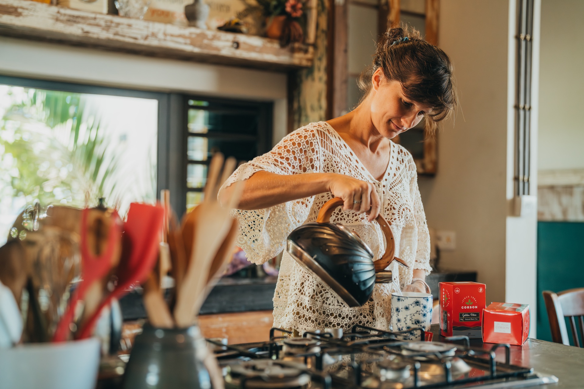 A woman pouring a kettle into a cup beside Corson tea boxes in a sunlit Mauritian kitchen during a morning tea ritual